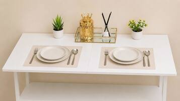 A white dining table with two sets of plates, cutlery, and napkins, flanked by small potted plants and a decorative golden item.