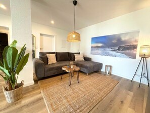 A living room with a grey sofa, a woven rug, a small round table, and a large beach-themed wall art. - Holiday Apartment With 1 Bedroom (Cuxhaven)
