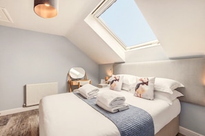 Ein Schlafzimmer mit schrägem Dach, einem Oberlicht, einem Bett mit weißer Bettwäsche, einem Nachttisch mit Lampe und einer Heizung.