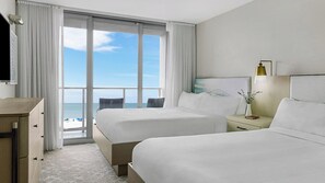 Habitación de hotel con dos camas, un escritorio, una mesita de noche y vista a la playa.