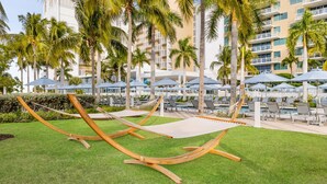 A hotel pool area with multiple lounge chairs and umbrellas, surrounded by palm trees and a building with balconies. - Marriott's Crystal Shores 2 Bedroom Villa With Full Resort Access (Marco Island)