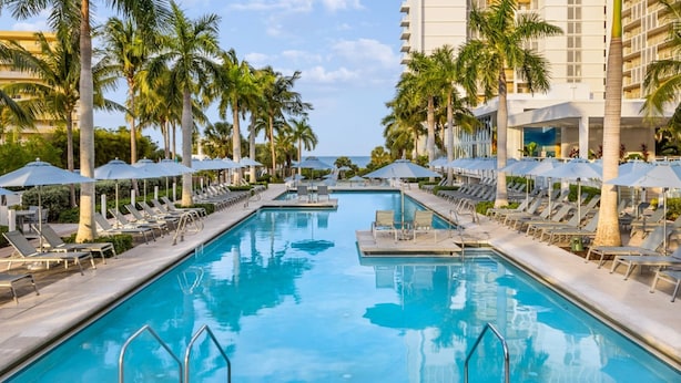 A swimming pool with lounge chairs and umbrellas, surrounded by palm trees and a hotel building. - Marriott's Crystal Shores 2 Bedroom Villa With Full Resort Access (Marco Island)