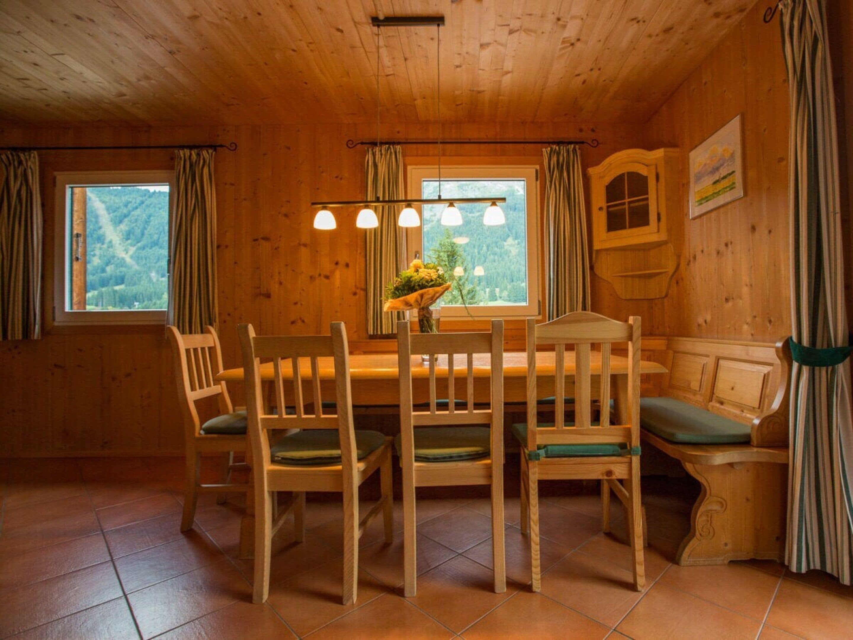 A wooden interior with a dining table, chairs, a bench, and a window with curtains.