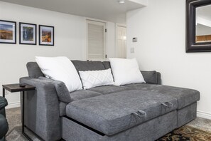 Ein graues Ecksofa mit weißen Kissen, ein kleiner Beistelltisch und ein gerahmtes Bild an der Wand.