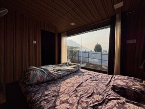 Un dormitori amb un llit, parets de fusta, una finestra amb vista a una muntanya i una tanca metàl·lica a l'exterior.