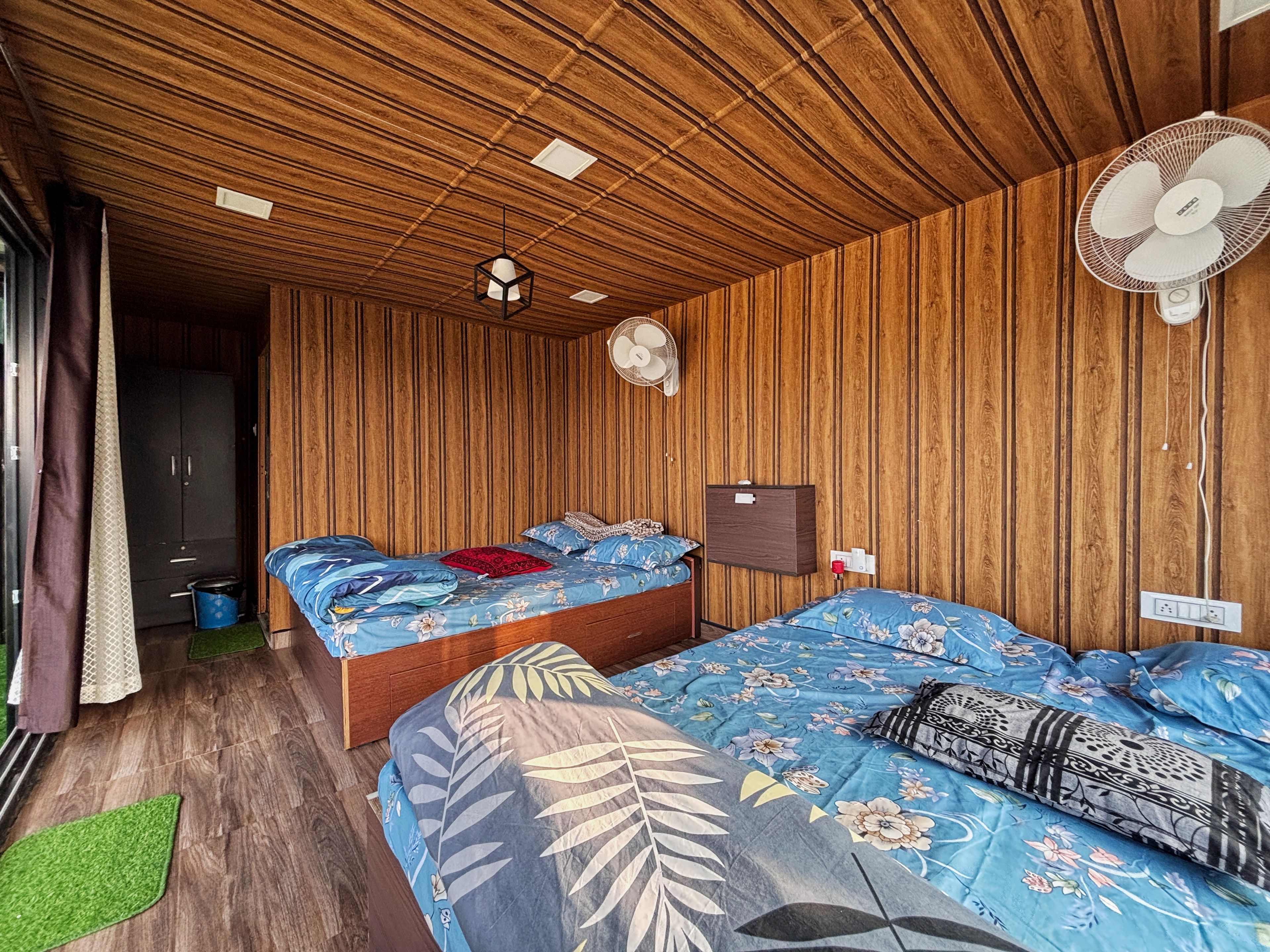 Una habitación con paneles de madera, una cama con ropa de cama floral, una mesita de noche, un ventilador de pared y una ventana con cortinas.