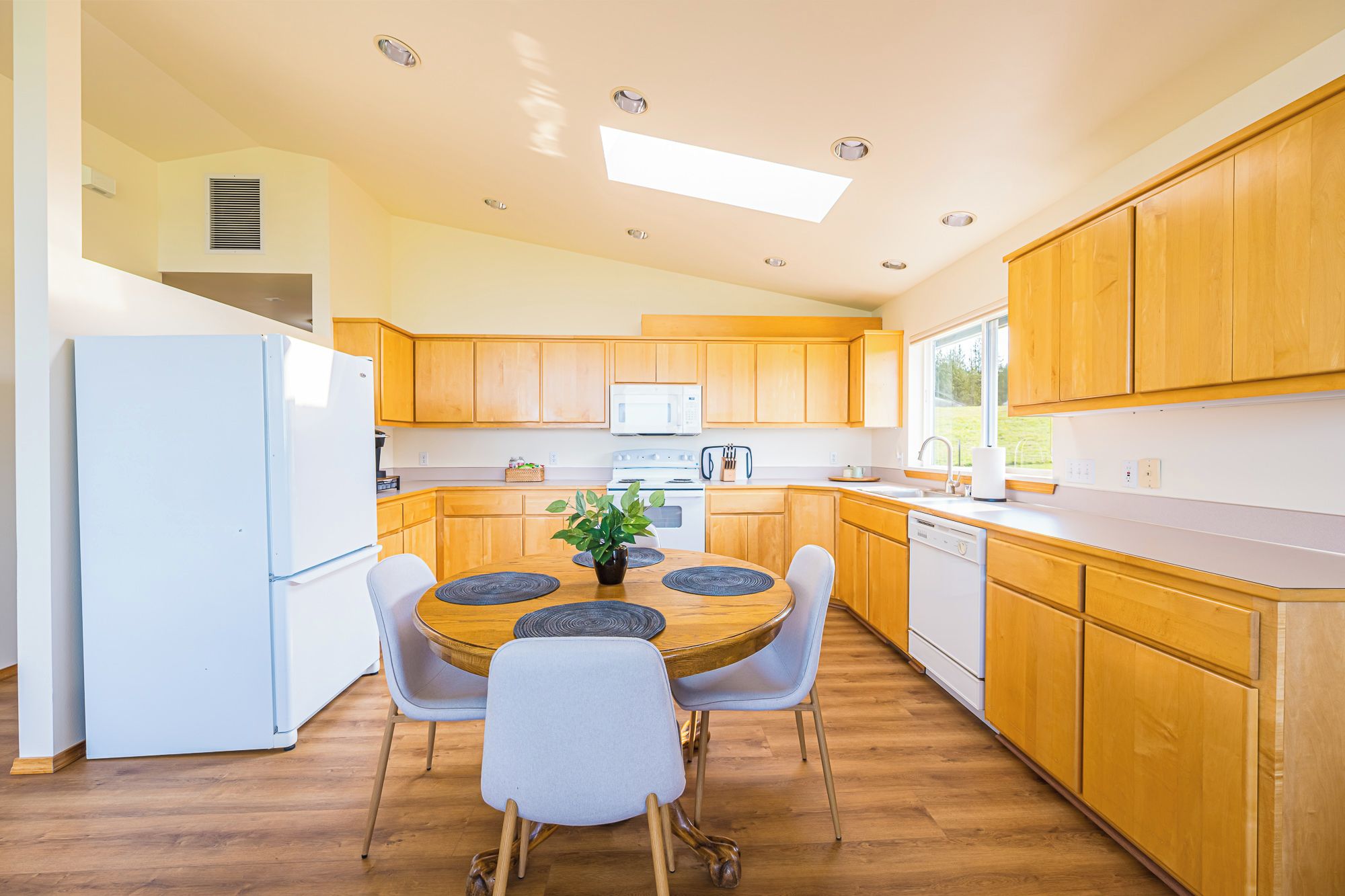 Une cuisine moderne avec des meubles en bois, un réfrigérateur blanc, une table à manger avec des chaises et une fenêtre qui laisse entrer la lumière naturelle.