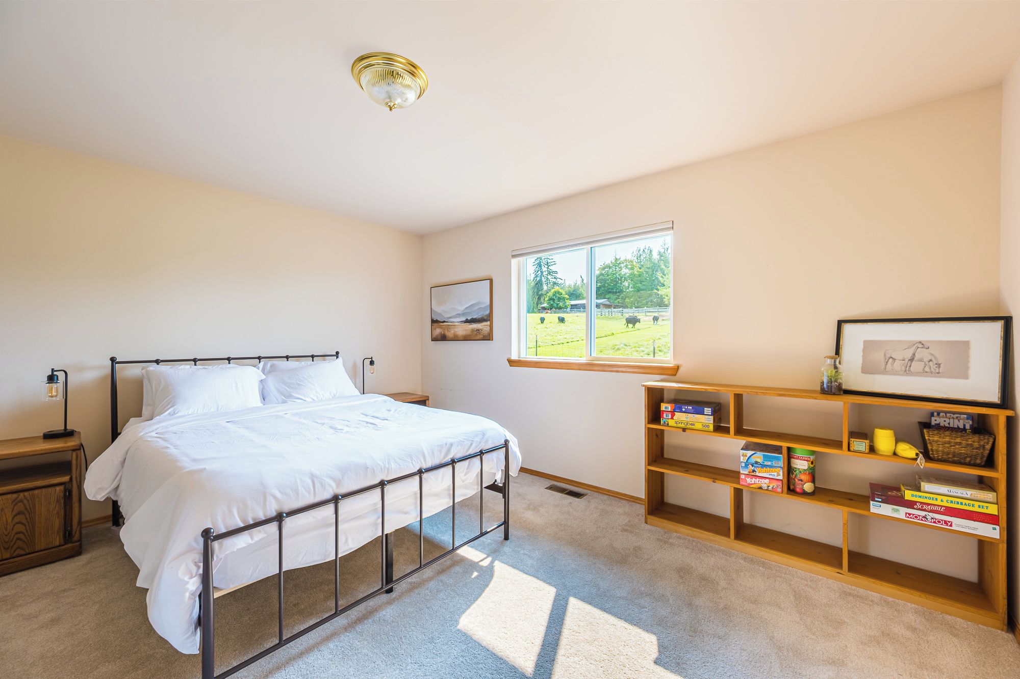 Une chambre à coucher avec un lit, une table de chevet, une fenêtre donnant sur une ferme et une bibliothèque remplie de livres et de jeux.