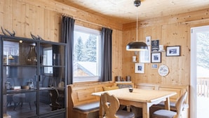 Interior de una cabaña de madera con una mesa de comedor, bancos, un armario y un ventanal grande.
