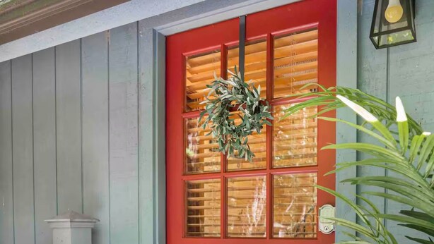 A dog sitting on a doormat with a wreath on a red door.