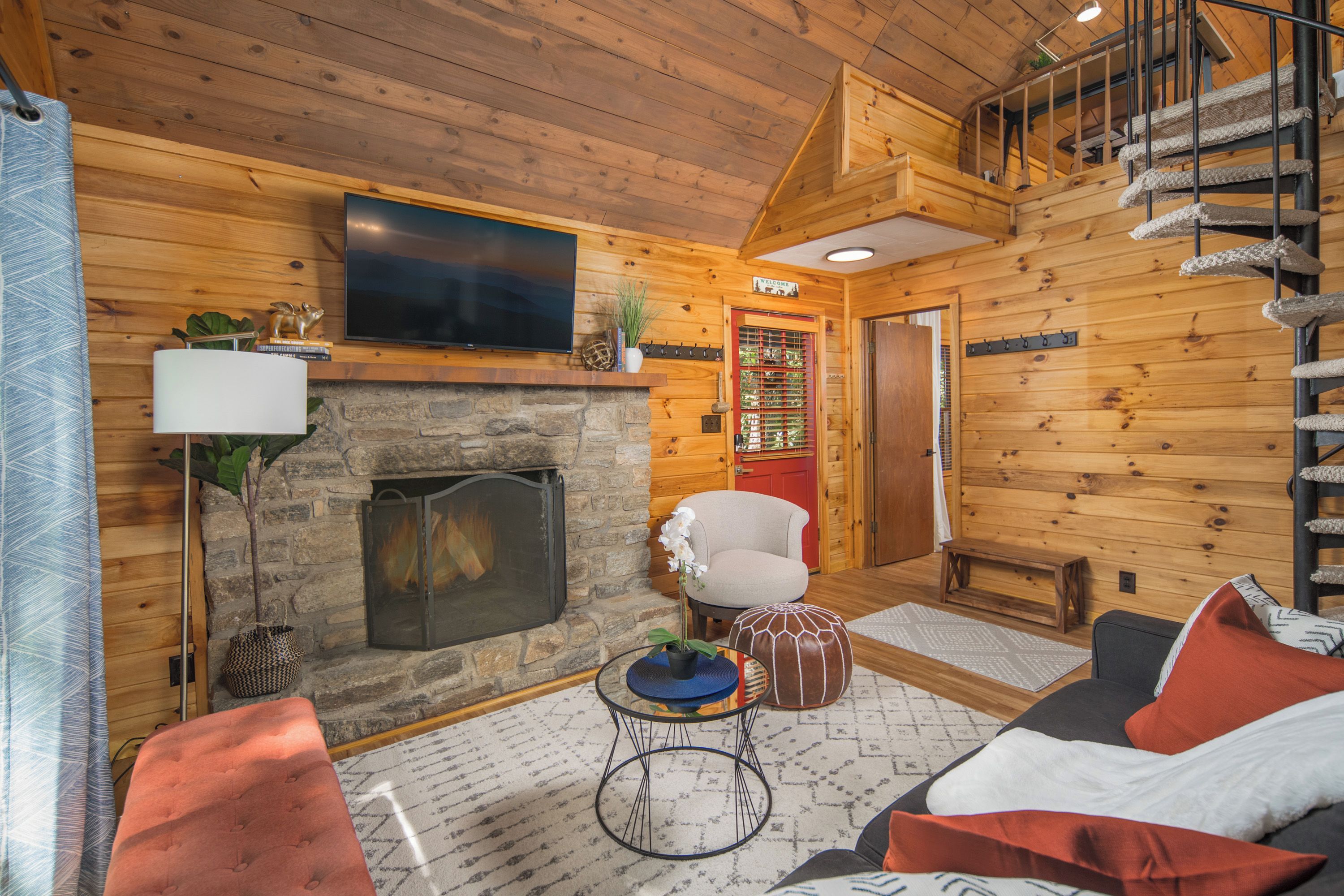 Ein gemütliches Blockhaus-Interieur mit Kamin aus Stein, einem darüber montierten Fernseher, einer bequemen Couch und einer Wendeltreppe.