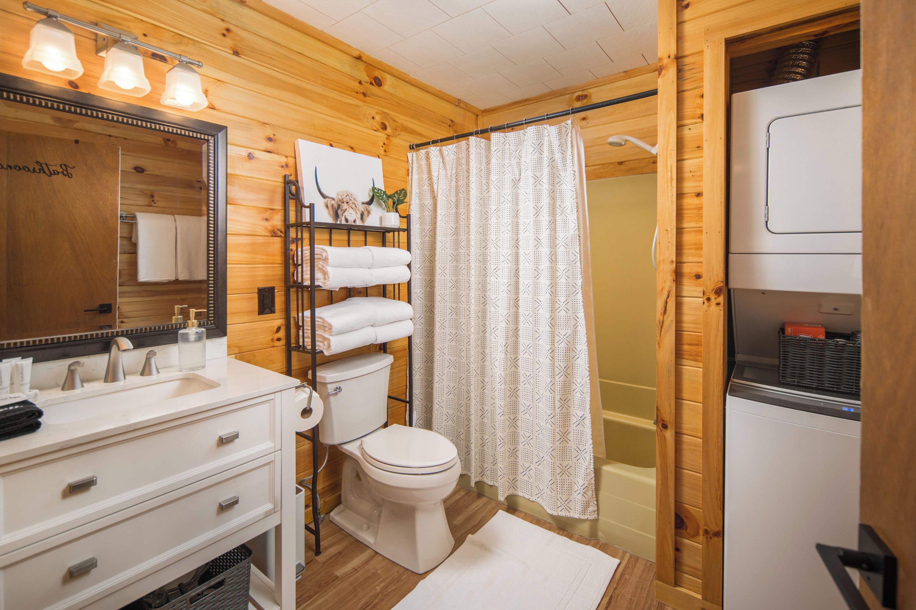 Baño de cabaña de madera con lavamanos blanco, espejo, inodoro y cortina de ducha.