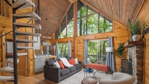 Interior espacioso de una cabaña con un altillo con cama, una sala de estar acogedora con sofá y reposapiés, un comedor y ventanas amplias que ofrecen vistas del bosque circundante.