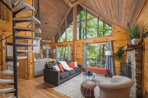 A spacious cabin interior with a loft bedroom, a cozy living area with a sofa and ottoman, a dining area, and large windows offering a view of the surrounding forest. - Hot Tub A-Frame Retreat, EV+Dogs OK, Blue Ridge Pkwy (Deep Gap)