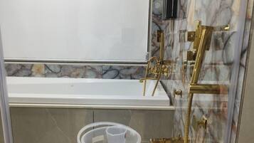 Un bany amb banyera, dutxa amb detalls daurats i un recipient de plàstic transparent a terra.