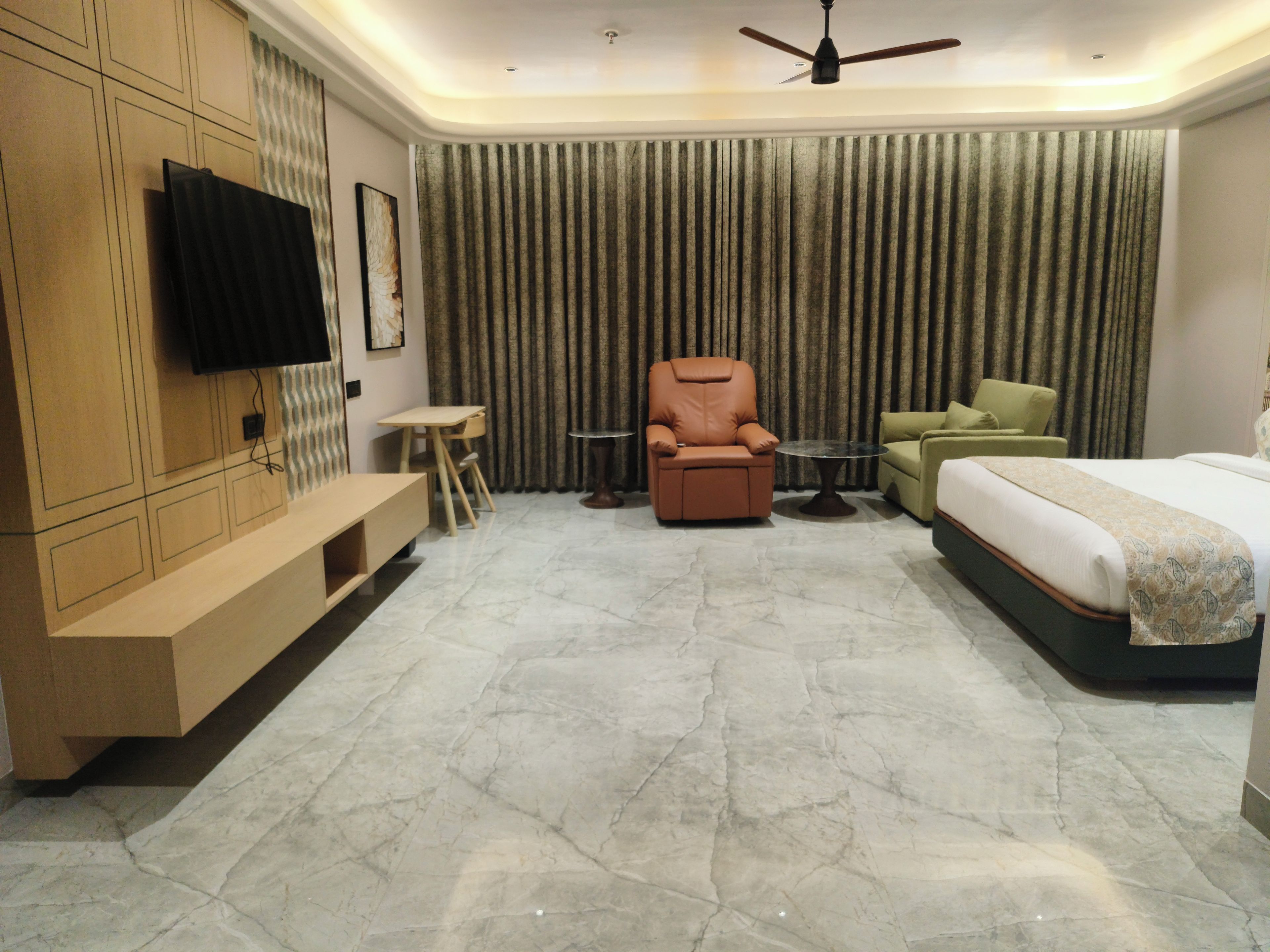 A modern hotel room with a flat-screen TV mounted on the wall, a sofa, a recliner, a green armchair, a small round table, and a bed with a patterned bedspread.