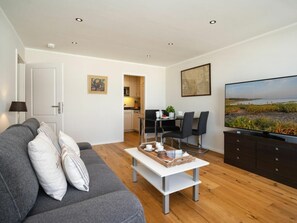 A modern living room with a sofa, a coffee table, a TV, and a dining area with chairs and a table. - Holiday Apartment With 1 Bedroom (Sylt)