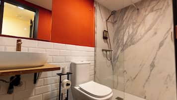 Un baño moderno con inodoro blanco, cabina de ducha con vidrio y lavamanos blanco sobre encimera de madera.