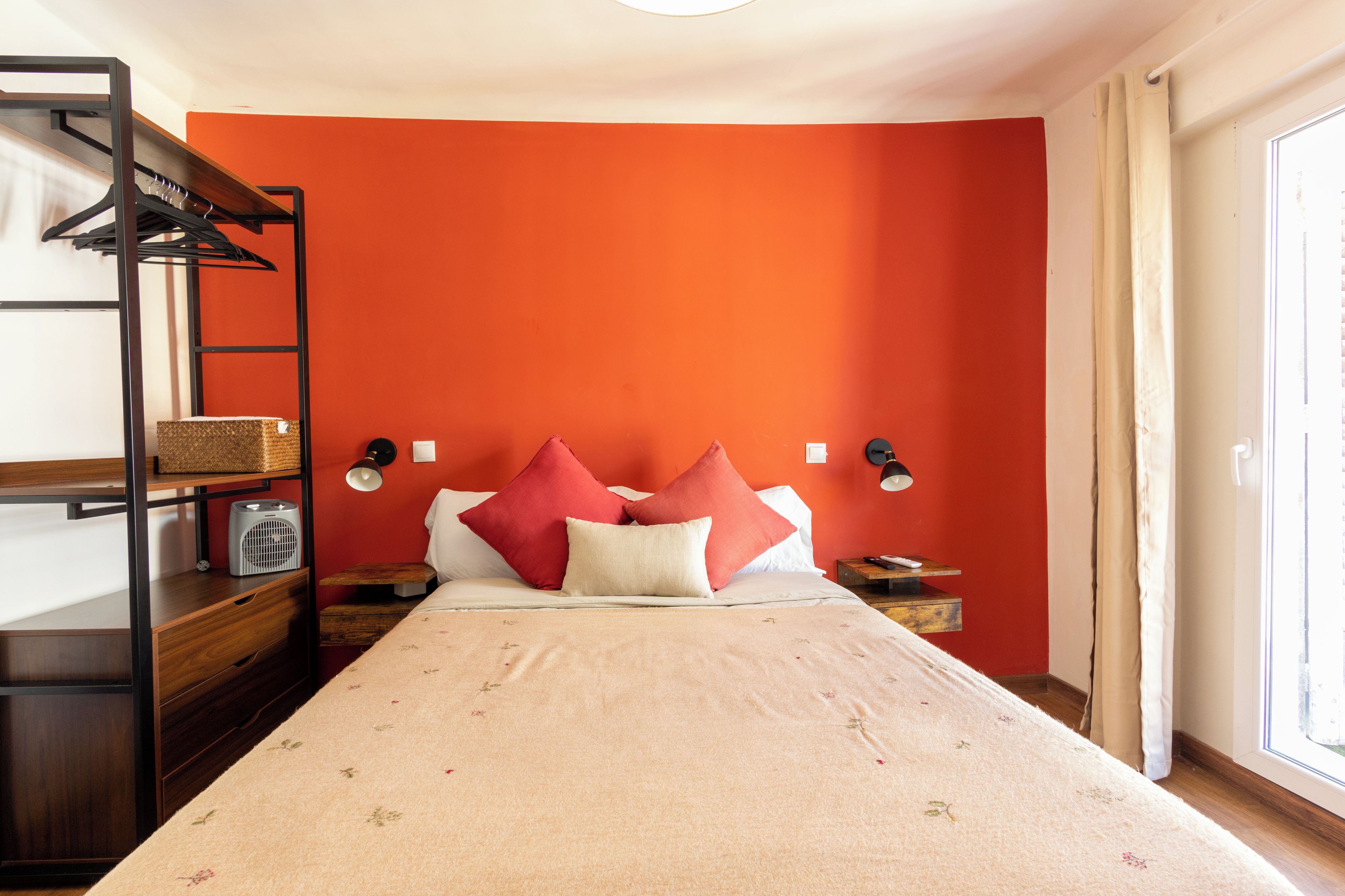 Una habitación con cama, mesita de noche, lámpara, estante con una cesta y ventana con cortinas.