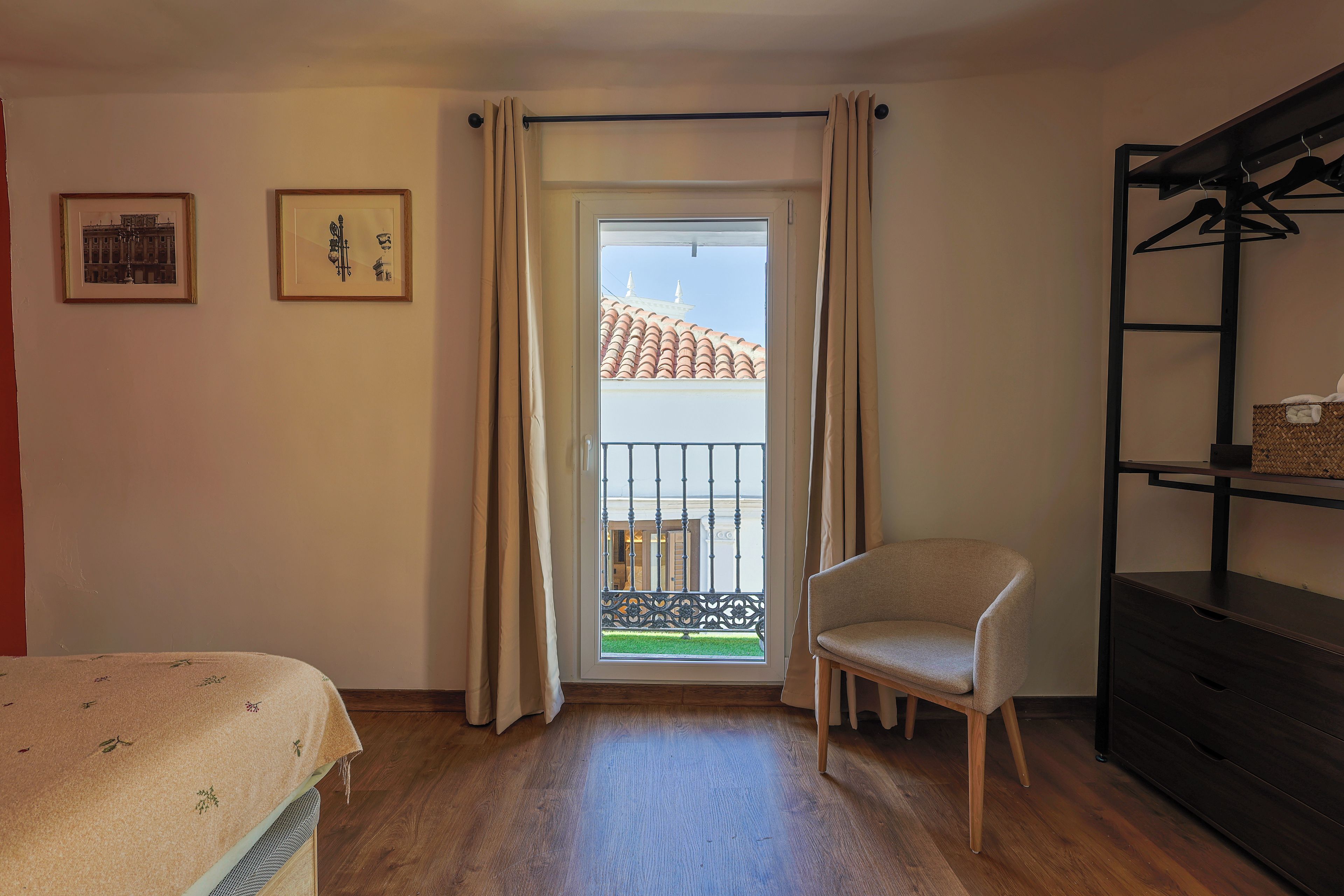 A room with a bed, a chair, a balcony with a railing, and two framed pictures on the wall.