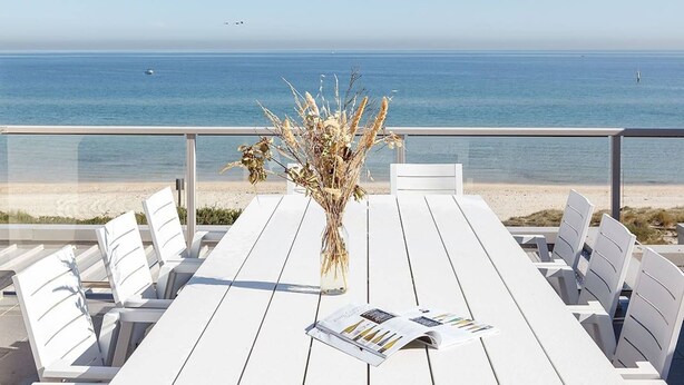 Una mesa de madera blanca con sillas y un jarrón de plantas secas, con vista a una playa y al océano.