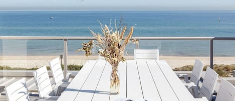 A white wooden table with chairs and a vase of dried plants, overlooking a beach with a clear view of the ocean.