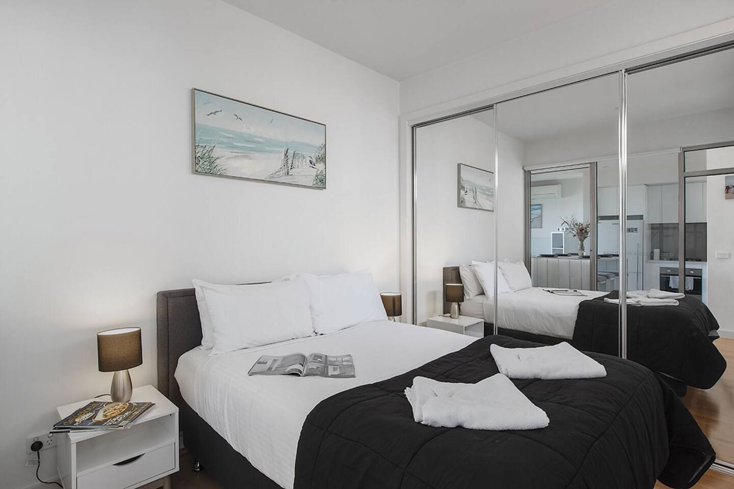 A modern hotel room with a large bed, a bedside table with a lamp, a magazine, and a painting on the wall.