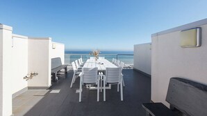 Una terraza en la azotea con una larga mesa de comedor y sillas, un rincón de descanso y vistas al océano.