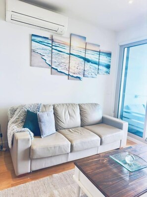 Un salón moderno con un sofá beige, una mesa de centro de cristal y un amplio ventanal con vistas al mar.