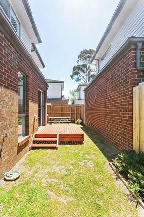 A backyard with a wooden deck, a brick wall, and a grassy area. - Hamptons Style Haven for Laidback Family Living (Rosebud)