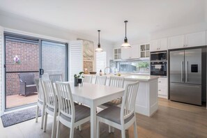 A modern kitchen with white cabinetry, a stainless steel refrigerator, and a dining area with white chairs. - Hamptons Style Haven for Laidback Family Living (Rosebud)