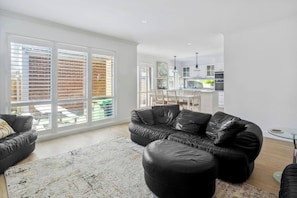 A spacious living room with a black leather sofa, a coffee table, and a large window with blinds. - Hamptons Style Haven for Laidback Family Living (Rosebud)