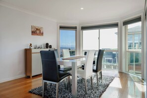 A dining area with a table and chairs, a view through large windows, and a small shelf with decorative items. - Beachfront at Bonbeach A Coastal Lifestyle Gem (Bonbeach)
