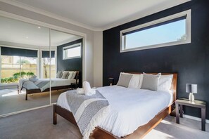 A modern bedroom with a large bed, a nightstand, a mirror, and a window. - Beachfront at Bonbeach A Coastal Lifestyle Gem (Bonbeach)