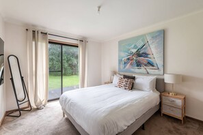 A bedroom with a large bed, a painting on the wall, a nightstand, and a window with curtains. - Solanders Retro Residence Calming Days by the Sea (Dromana)