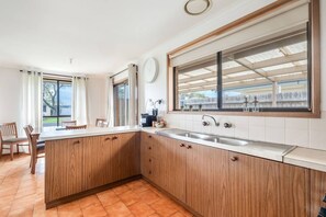 Een keuken met houten kastjes, een groot raam en een eethoek met stoelen.