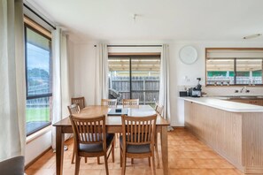 Een eetruimte met een houten tafel en stoelen, een keuken met een spoelbak en aanrecht, en grote ramen met gordijnen.