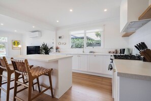 A modern kitchen with white cabinetry, a central island, wooden chairs, and a built-in stove. - Hamptons on Heales Classic Beach House Charm (Dromana)