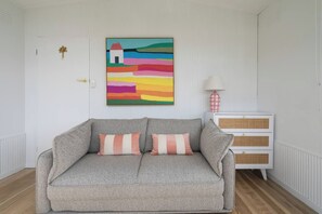 Una habitació amb un sofà gris, dos almofadons amb ratlles, un quadre abstracte i colorit, una taula de nit blanca amb una làmpada, i un objecte decoratiu petit a la paret.