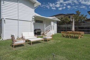 Una casa blanca amb una zona de terrassa que compta amb un sofà blanc, dues cadires, una banca de fusta i una barbacoa.