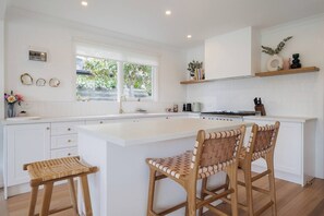 A modern kitchen with white cabinetry, a large island with bar stools, a window with greenery outside, and a shelf with decorative items. - Hamptons on Heales Classic Beach House Charm (Dromana)