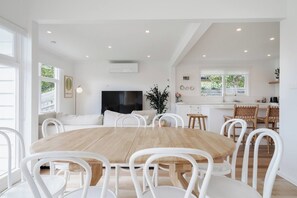 A spacious dining area with a wooden table and white chairs, a kitchen with white cabinets and a window, and a flat-screen TV mounted on the wall. - Hamptons on Heales Classic Beach House Charm (Dromana)