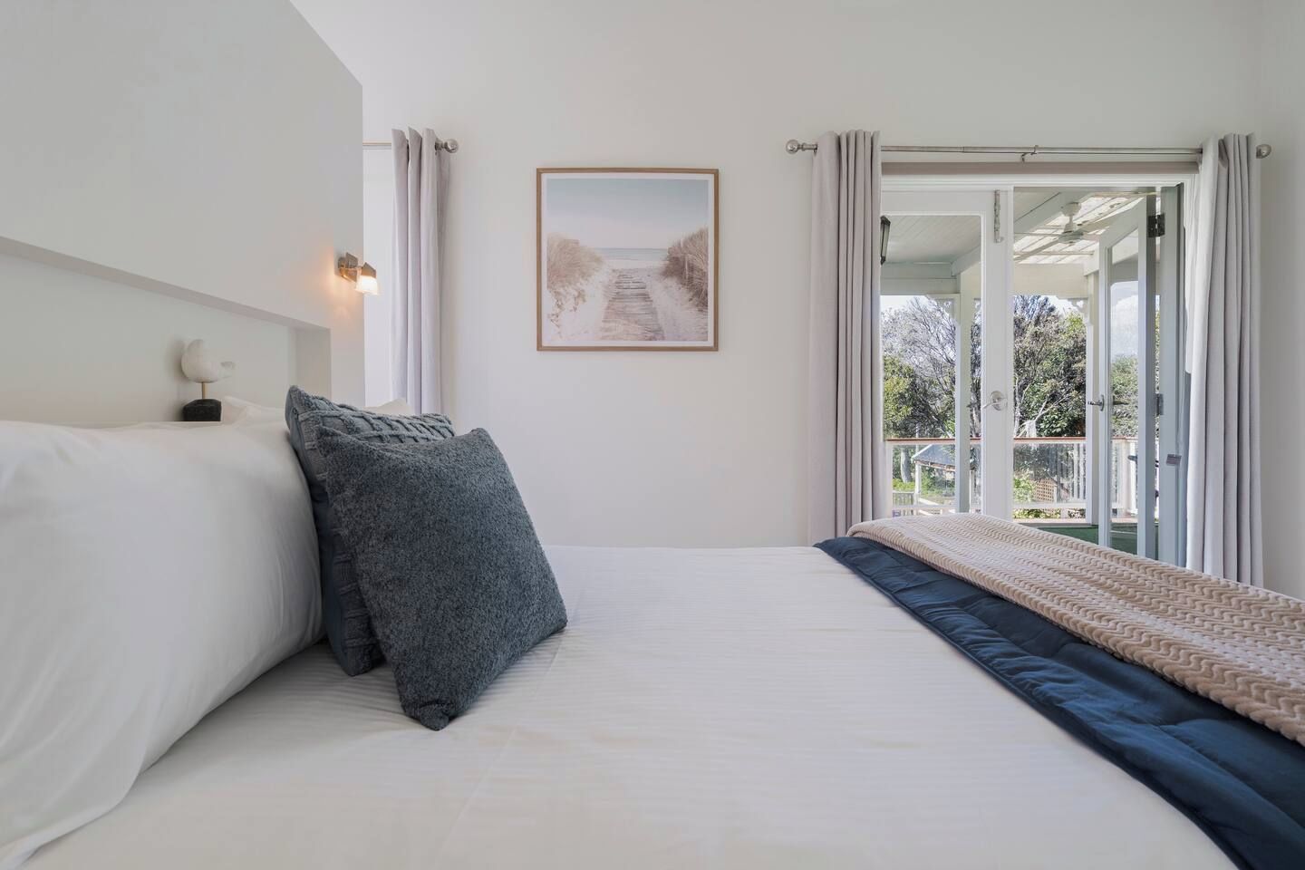 Une chambre à coucher avec un lit, des oreillers, un tableau encadré au mur et une vue sur l’extérieur, visible à travers une porte ouverte.