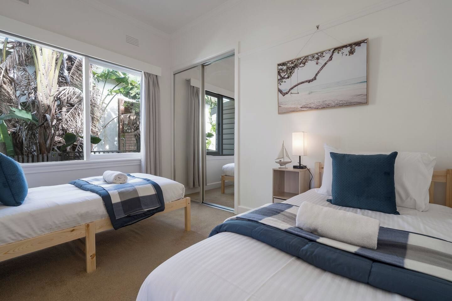 A hotel room with two beds, a window with a view of greenery, a painting on the wall, and a bedside table with a lamp.