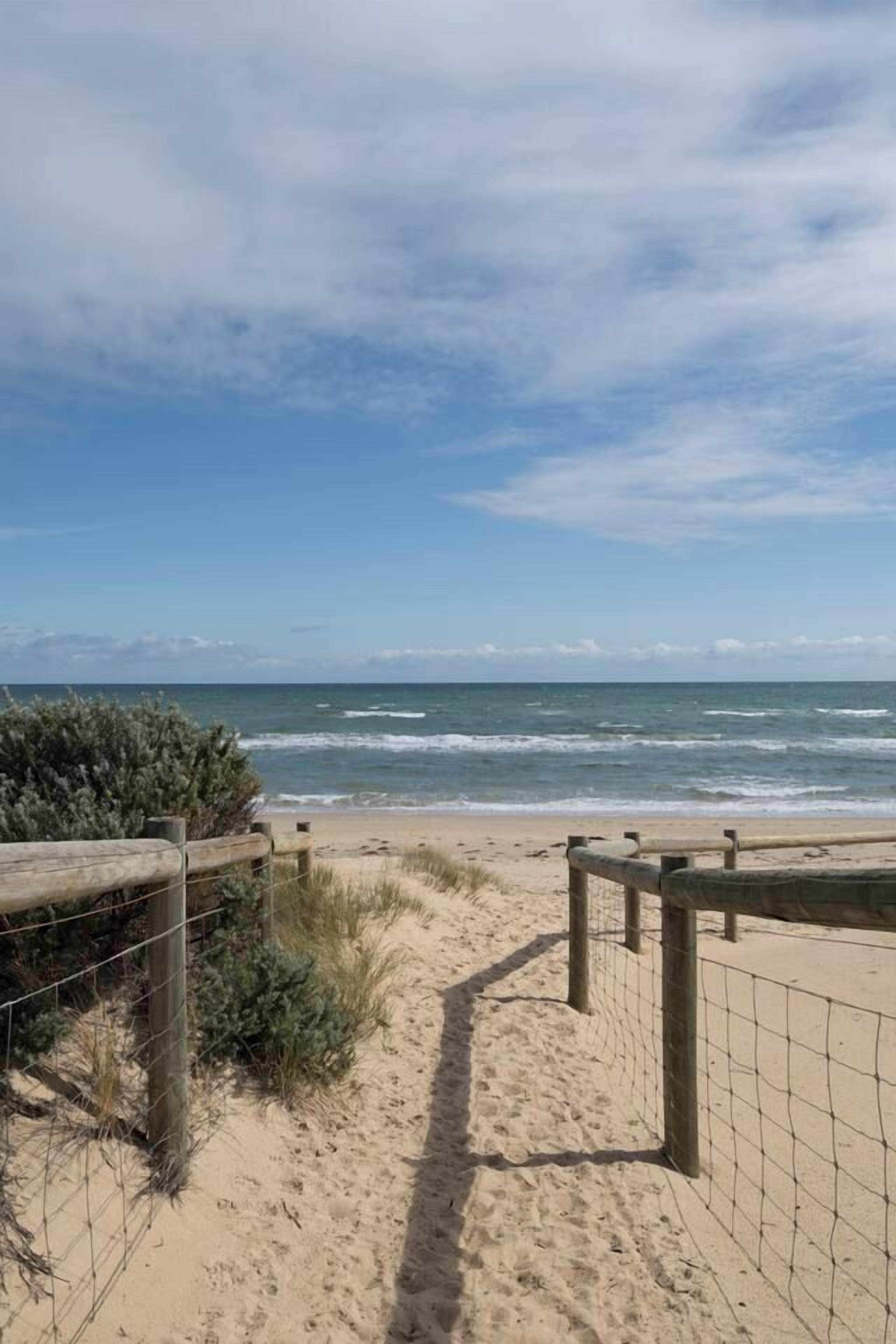 A sandy beach path leading to the ocean with a wooden fence and wire mesh on the left.
