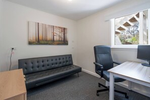 Ein Zimmer mit einer schwarzen Ledersofa, einem schwarzen Bürostuhl, einem weißen Schreibtisch mit Computerbildschirm und einem Waldgemälde an der Wand.