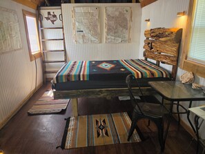 Une petite pièce comprenant un lit, une table, une chaise, une échelle et un mur orné de cartes.