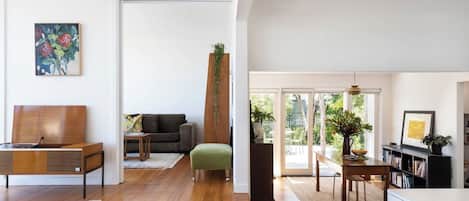 A spacious living area with a wooden floor, a sofa, a coffee table, and a dining table with chairs. There is a bookshelf and a plant on the right side.