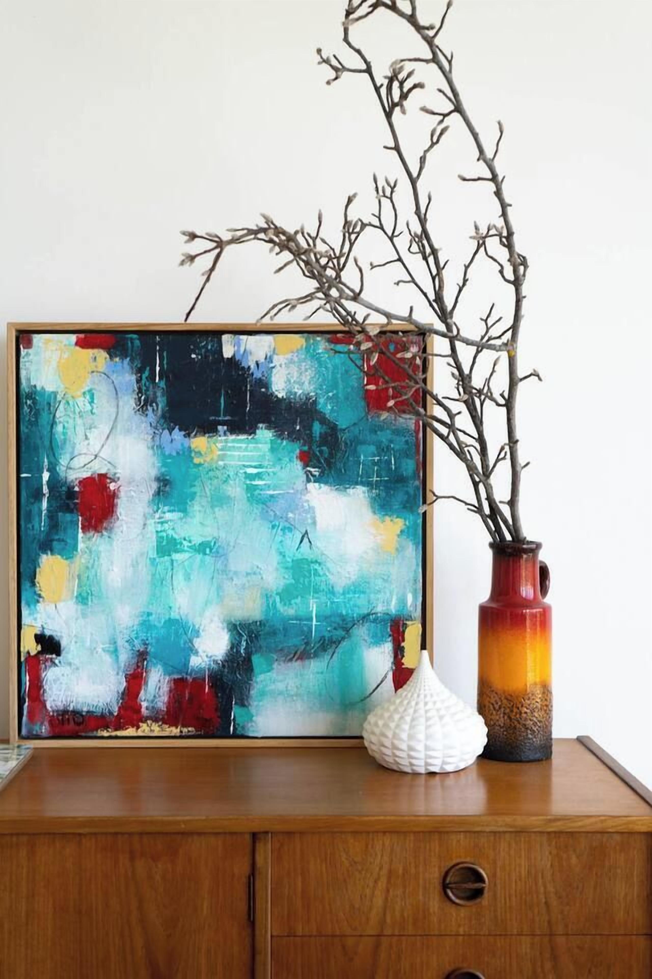A framed abstract painting on a wooden dresser with a vase and a decorative object beside it.