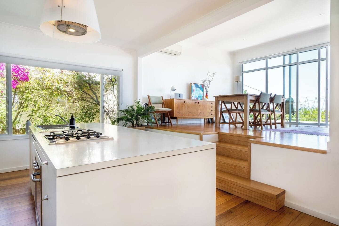 A modern kitchen with a central island, a dining area with a wooden table and chairs, and large windows offering a view of greenery.